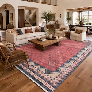 May include: A large rectangular area rug featuring a faded red center with a repeating diamond pattern, bordered by a wide navy blue and cream geometric zigzag frame with a fringed edge, covering a significant portion of the wooden floor.