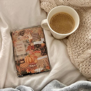 以下が含まれることがあります： カボチャ、松ぼっくり、リンゴのバスケット、そして「leaves books lattes」、「this is my emotional support kindle」、「autumn days」、「one more chapter」という文字が描かれた秋をテーマにしたコラージュ付きの透明な電子書籍リーダーケース。コーヒーマグの隣に配置されています。