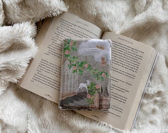 May include: A clear rectangular phone case featuring a decorative collage of a white stag, pink roses, green vines, a stone archway, a small golden crown, and the text "She's an assassin" and "I will defeat you" resting on an open hardcover book.