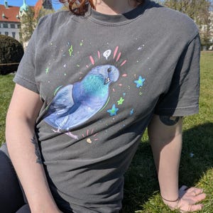 May include: Dark gray t-shirt featuring a blue and teal pigeon illustration with a pink halo and question mark. The design includes colorful stars and sparkles. The shirt is worn outdoors on a sunny day.