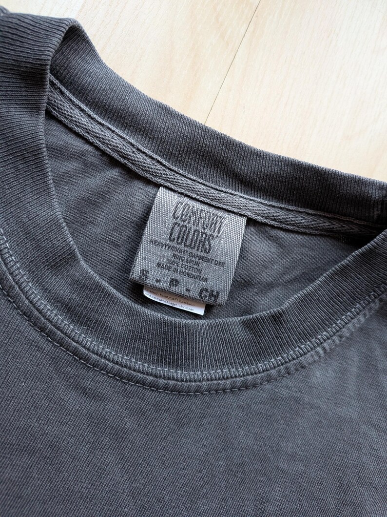May include: Close-up of a charcoal gray t-shirt collar and tag. The tag reads "COMFORT COLORS" and includes text about the fabric content and origin. The shirt appears to be made of a soft, heavyweight material.
