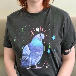 May include: Dark gray t-shirt featuring a cartoon pigeon with blue and purple feathers, surrounded by stars and sparkles. The pigeon has a question mark above its head. The shirt is a crew neck with short sleeves.