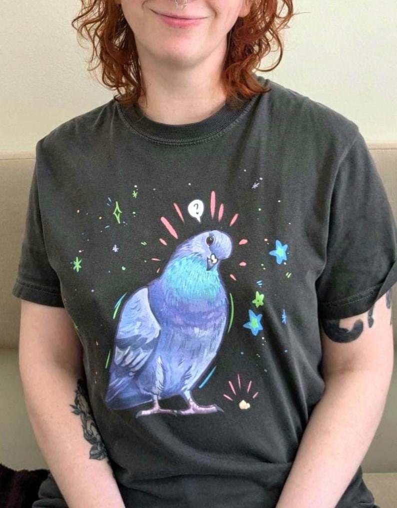 May include: Dark gray t-shirt featuring a cartoon pigeon with blue and purple feathers, surrounded by stars and sparkles. The pigeon has a question mark above its head. The shirt is a crew neck with short sleeves.