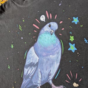May include: Dark grey t-shirt featuring a whimsical illustration of a blue and teal pigeon with a question mark above its head. The design includes colorful stars and sparkles. The shirt is made of a soft material.