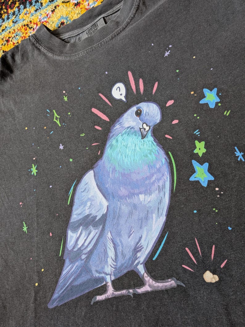 May include: Dark grey t-shirt featuring a whimsical illustration of a blue and teal pigeon with a question mark above its head. The design includes colorful stars and sparkles. The shirt is made of a soft material.