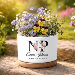 May include: A white ceramic planter with a wooden base, filled with colorful flowers and small stones. The planter features the initials "NP" with floral accents, the name "Emma Johnson", and the title "Nurse Practitioner".