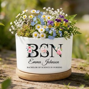 May include: A white ceramic planter with the letters "BSN" and the name "Emma Johnson" in black script, filled with a variety of colorful flowers. The planter sits on a wooden base, perfect for a desk or windowsill.