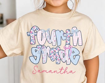 May include: A cream-colored short-sleeve t-shirt featuring the text "Fourth Grade" in a multi-patterned font with blue, pink, and floral designs, accented by small bows. The name "Samantha" is printed in pink script below the main text.
