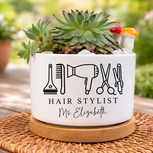 May include: White ceramic planter with a succulent plant, featuring black line art of hair styling tools and the text "HAIR STYLIST Ms. Elizabeth". The planter sits on a round, woven coaster.
