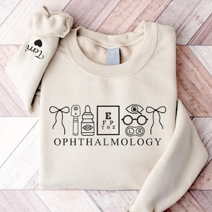May include: A beige sweatshirt with black ophthalmology-themed graphics, including eye charts, glasses, and eye care tools. The word "OPHTHALMOLOGY" is printed below the graphics. The left sleeve has the name "Terri" and a heart.