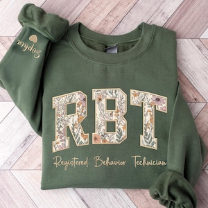 May include: An olive green crewneck sweatshirt featuring the letters RBT in a floral print with a gold outline. Below the letters, the text Registered Behavior Technician is printed in gold. The name Sophia and a small heart are on the sleeve.