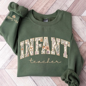 May include: An olive green crewneck sweatshirt featuring the word INFANT in large, block-style letters filled with a floral pattern, and the word teacher in cursive script below. The sleeve displays the text Mrs. Smith with a small heart icon.