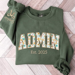 May include: A forest green sweatshirt with the word "ADMIN" in a floral design. The text "Est. 2025" is below the word "ADMIN". The left sleeve has the name "Sophia" embroidered in gold with a bow design.