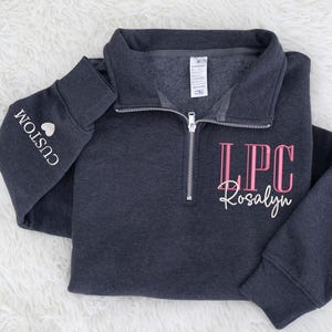 May include: A dark charcoal gray quarter-zip sweatshirt featuring a silver zipper and a collar. The front displays the pink text LPC above the white script name Rosalyn. The left sleeve features the white text CUSTOM with a heart icon.