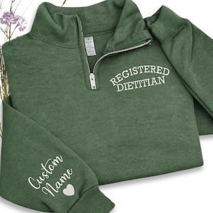 May include: A green quarter-zip sweatshirt with the words "REGISTERED DIETITIAN" in white script on the chest. The left sleeve has "Custom Name" and a heart in white script. The sweatshirt is folded on a white surface.