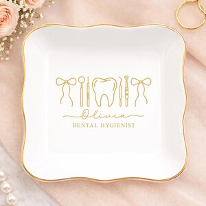 Könnte beinhalten: Eine weiße Keramik-Schmuckschale mit goldenem Rand und gewelltem Rand. Die Schale zeigt eine goldene Umrisszeichnung von zahnärztlichen Instrumenten, einem Zahn und dem Namen "Olivia" mit "Dental Hygienist" darunter.