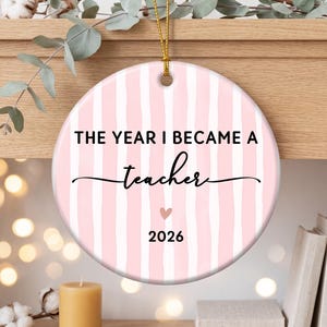 May include: A round ceramic ornament featuring a light pink and white vertical striped pattern. The text reads THE YEAR I BECAME A teacher in black lettering, with a small pink heart icon above the year 2026. A gold string is attached at the top.