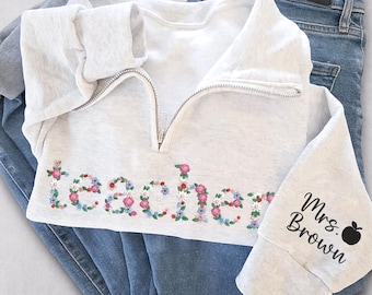 May include: A light gray sweatshirt with a half-zip neckline and the word "teacher" spelled out in colorful floral embroidery. The right sleeve has the text "Mrs. Brown" and a black apple graphic. The sweatshirt is paired with blue jeans.