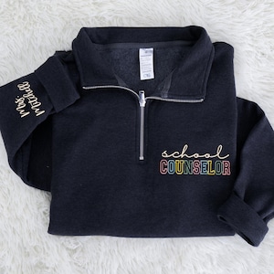 May include: A dark navy blue quarter-zip sweatshirt with the words "school COUNSELOR" embroidered in rainbow colors on the front. The left sleeve has the embroidered text "Mrs. Mitchell" in a cursive font.