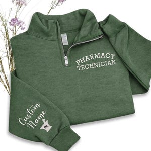 May include: A forest green quarter-zip sweatshirt with the words "PHARMACY TECHNICIAN" in white script on the chest. The left sleeve features "Custom Name" and a mortar and pestle graphic. The sweatshirt has a ribbed cuff and collar.