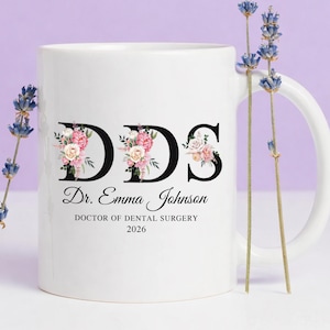 May include: White ceramic mug with the letters "DDS" in black, each letter adorned with pink and white floral arrangements. The mug also features the name "Dr. Emma Johnson" and "DOCTOR OF DENTAL SURGERY 2026".