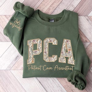 May include: An olive green sweatshirt with the letters "PCA" in a floral design. The words "Patient Care Assistant" are below the letters. The sleeve has the name "Amelia" and a pair of scissors embroidered in gold.