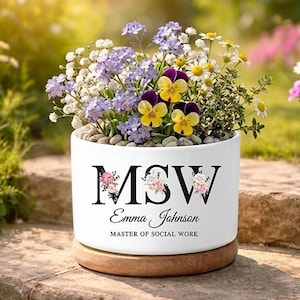 May include: A white ceramic planter with the initials "MSW" and the name "Emma Johnson" in black lettering, filled with a variety of colorful flowers. The planter sits on a wooden base, with a garden background.