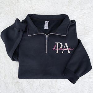 May include: A black quarter-zip sweatshirt with the letters "PA" embroidered in white and the word "drama" in pink script. The sweatshirt has a collar and a silver zipper. The sleeves are cuffed.