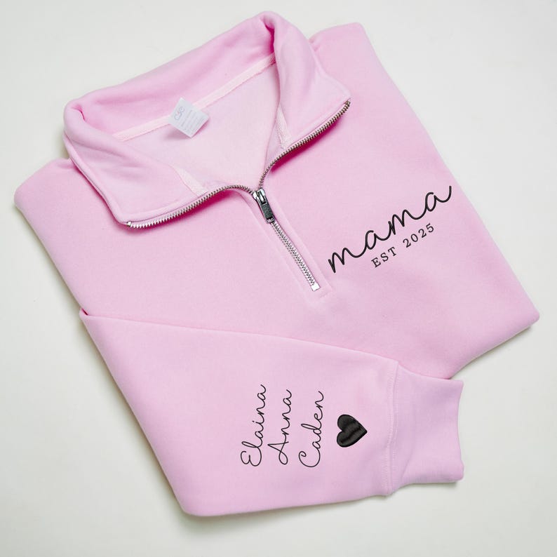 May include: A light pink sweatshirt with a quarter-zip collar. The word "mama" and "EST 2025" are printed in black script on the front. The sleeve features the names "Elaina, Anna, Caden" and a black heart.