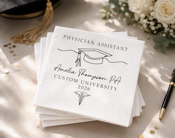 May include: A stack of white paper napkins with black text and a graduation cap illustration. The text reads "PHYSICIAN ASSISTANT" and includes a name, university, and the year 2026. A caduceus symbol is also present.
