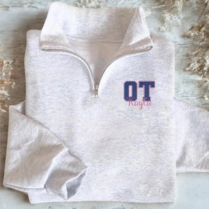 May include: A light gray sweatshirt with a quarter-zip neckline. The sweatshirt features the letters "OT" in a blue and pink design, with the name "Kayla" below. The sweatshirt is made of a soft material and has long sleeves.