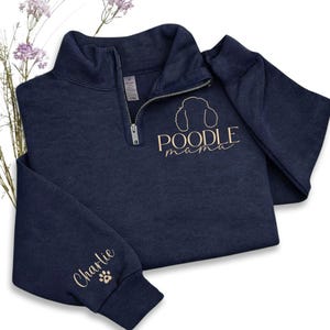 May include: Navy blue quarter-zip sweatshirt with the text "POODLE mama" and a poodle silhouette in gold. The sleeve features the name "Charlie" and a paw print. The sweatshirt is made of a soft material.