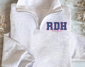May include: A light gray quarter-zip sweatshirt with the letters "RDH" in blue and pink, with the name "Kayla" underneath. The sweatshirt has a silver zipper and a folded collar.