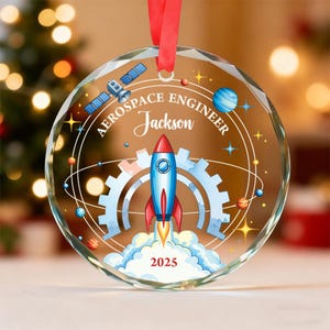 May include: A clear, round Christmas ornament with a red ribbon. The ornament features a rocket ship design with the text "AEROSPACE ENGINEER Jackson 2025". The design includes a satellite, planets, and gears.