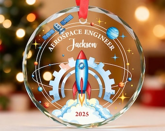 May include: A clear, round Christmas ornament with a red ribbon. The ornament features a rocket ship design with the text "AEROSPACE ENGINEER Jackson 2025". The design includes a satellite, planets, and gears.
