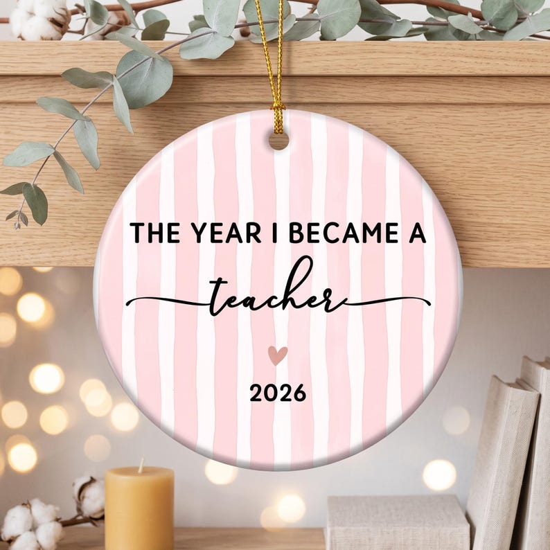 May include: A round ceramic ornament featuring a light pink and white vertical striped pattern. The text reads THE YEAR I BECAME A teacher in black lettering, with a small pink heart icon above the year 2026. A gold string is attached at the top.