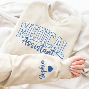 May include: A beige crewneck sweatshirt featuring the text MEDICAL ASSISTANT in a blue and white gingham pattern. The sleeve displays the name Sophia in blue cursive script followed by a small blue heart icon, all printed on the fabric.