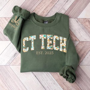 May include: A dark green sweatshirt with the text "CT TECH" in a floral design, and "EST. 2025" below. The left sleeve has the name "Sophia" embroidered with a bow and scissors design.