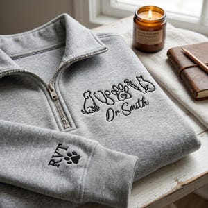 May include: A light gray quarter-zip sweatshirt featuring black embroidery on the chest with icons of a dog, stethoscope, paw print, syringe, and cat, followed by the text Dr. Smith. The sleeve cuff displays the embroidered text RVT and a paw print.