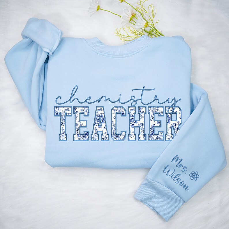 May include: A pale blue sweatshirt featuring the words "chemistry TEACHER" in a blue floral pattern. The sleeve displays "Mrs. Wilson" and a science symbol. The top is a crew neck style.