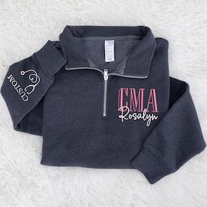 May include: A dark gray quarter-zip sweatshirt with a white stethoscope graphic and the word "CUSTOM" on the left sleeve. The front features the letters "CMA" in pink and the name "Rosalyn" in white.