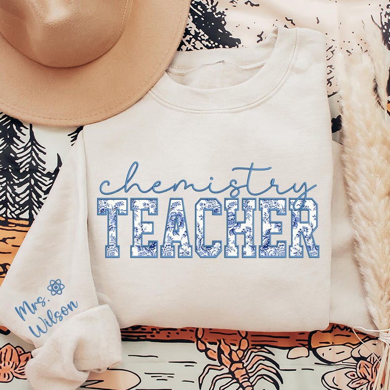 May include: A cream-coloured sweatshirt featuring the words "chemistry TEACHER" in blue script and block letters. The block letters have a blue floral pattern. The sleeve displays the name "Mrs. Wilson" and a science symbol in blue.
