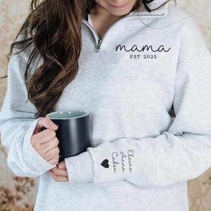 May include: A light gray sweatshirt with a quarter-zip neckline. The word "mama" and "EST 2025" are printed on the front. The sleeve features the names "Elaina, Anna, Caden" and a heart. A black mug is held in the hands.