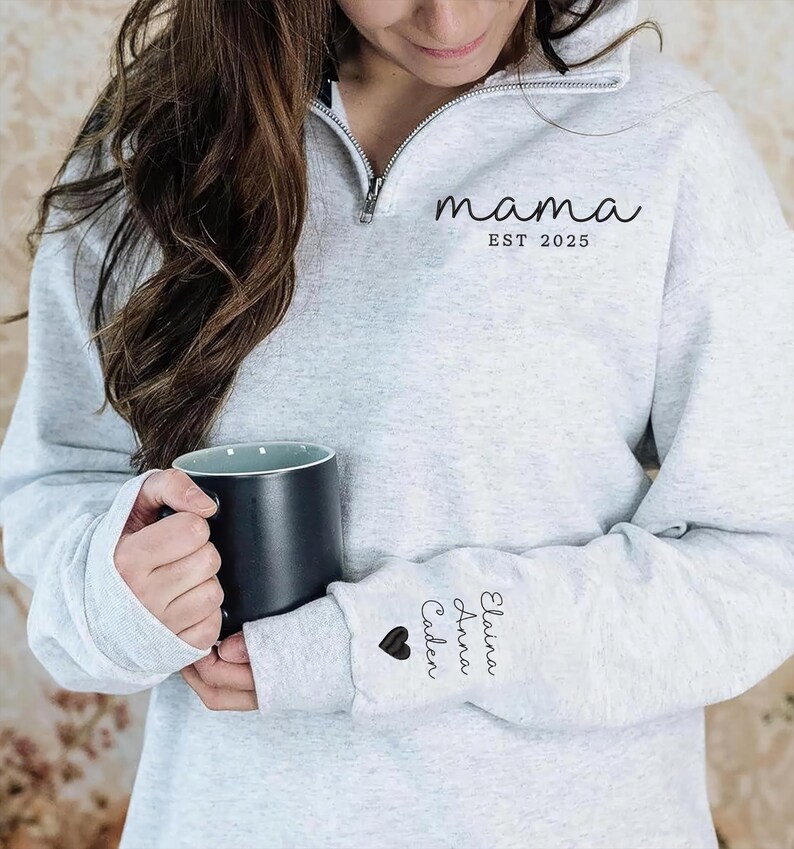 May include: A light gray sweatshirt with a quarter-zip neckline. The word "mama" and "EST 2025" are printed on the front. The sleeve features the names "Elaina, Anna, Caden" and a heart. A black mug is held in the hands.