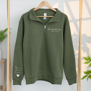 May include: Olive green sweatshirt with a quarter-zip collar. The word "mama" and "EST 2025" are embroidered on the chest. The left sleeve features the names "Elaina, Anna, Caden" and a heart symbol.