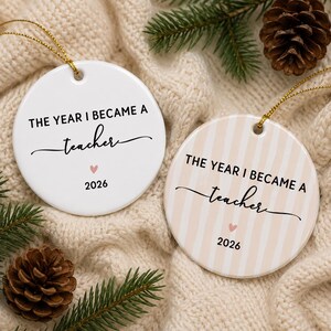 May include: Two round ceramic ornaments with gold hanging strings. One is solid white, the other has beige vertical stripes. Both feature the black text THE YEAR I BECAME A teacher, a small pink heart, and the year 2026 printed on the front surface.