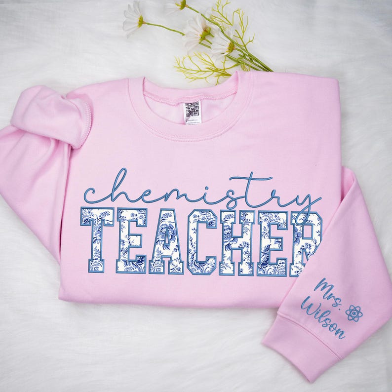 May include: A pale pink Chemistry Teacher sweatshirt. The word "chemistry" is written in a cursive blue font above the word "TEACHER", which is in a blue floral pattern. The right sleeve has the name "Mrs. Wilson" and a chemistry symbol.