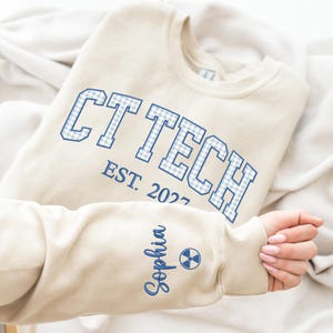 May include: A beige crewneck sweatshirt featuring the text CT TECH in blue and white gingham block letters, with EST. 2027 printed below. The sleeve is embroidered with the name Sophia in blue script next to a blue radiation symbol icon.