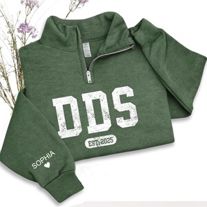 May include: A green quarter-zip sweatshirt with the letters "DDS" in distressed white lettering. Below the letters is "EST. 2025". The sleeve cuff displays the name "Sophia" and a heart symbol. The sweatshirt is folded.