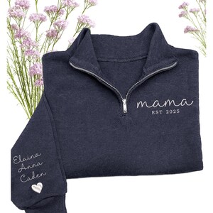 May include: A navy blue quarter-zip sweatshirt with the word "mama" and "EST 2025" embroidered in white. The left sleeve has the names "Elaina, Anna, Caden" and a heart embroidered in white. The sweatshirt has a silver zipper.